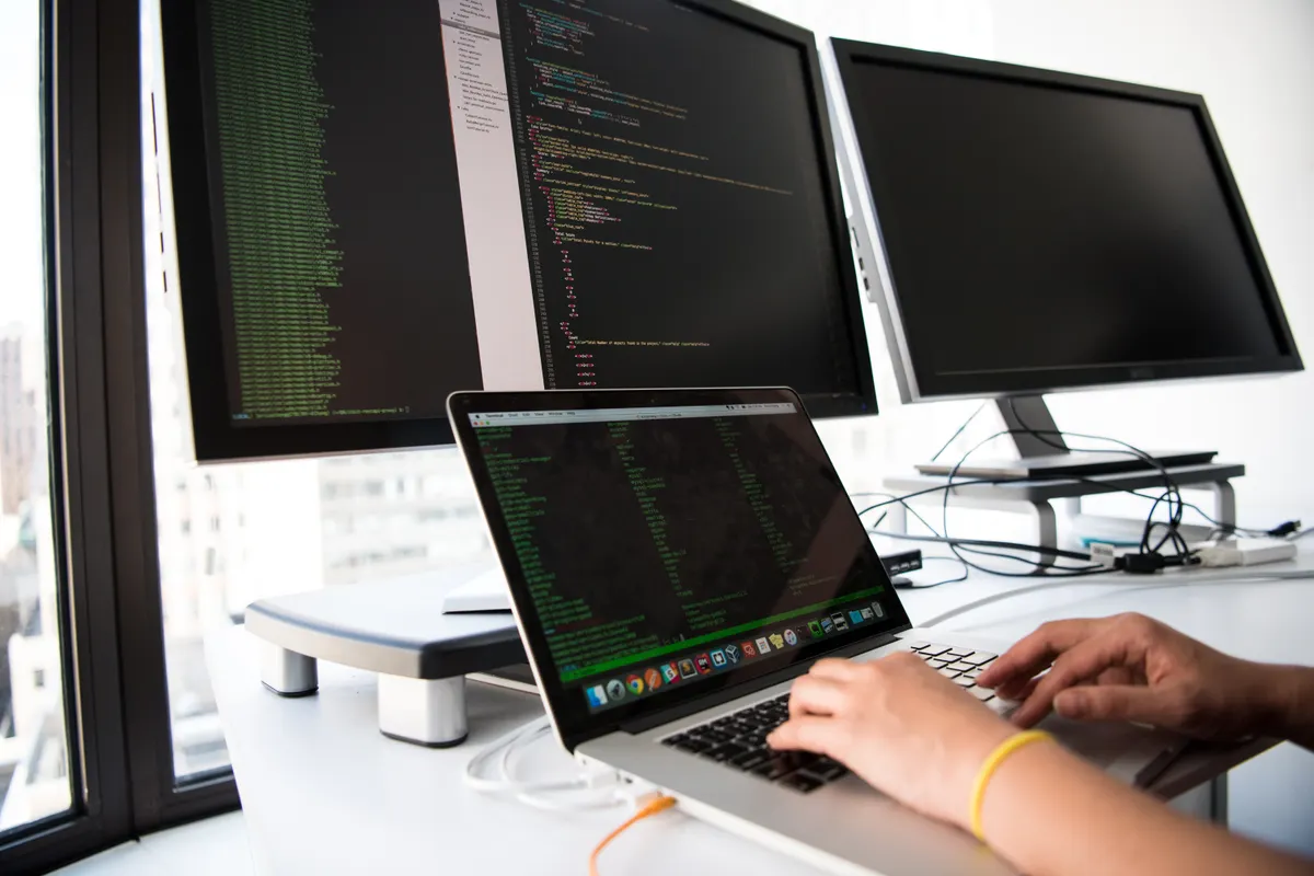 Tangan mengetik kode di laptop dengan dua monitor di meja kerja